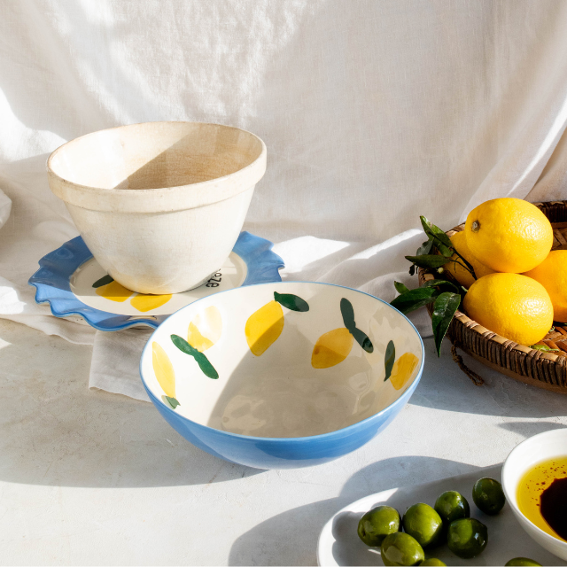 Decorative ceramic bowl with lemon design on a white surface with lemons and olives. Sun shining through with white fabric in background.