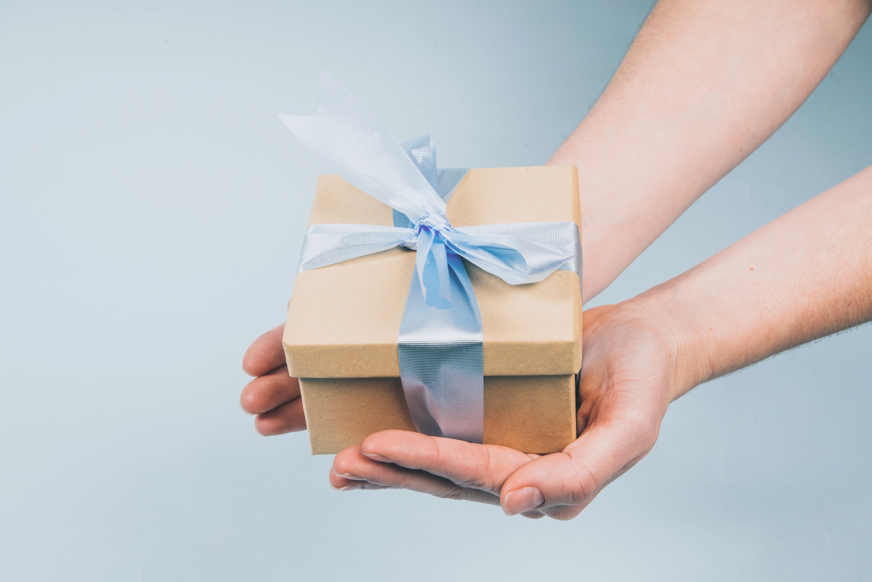 Two hands coming from outside the photo holding a gift box with baby blue ribbon tied around it on a light blue gradient background