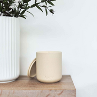 Beige ceramic Aery candle on a wooden shelf with a white vase and green plant in the background.