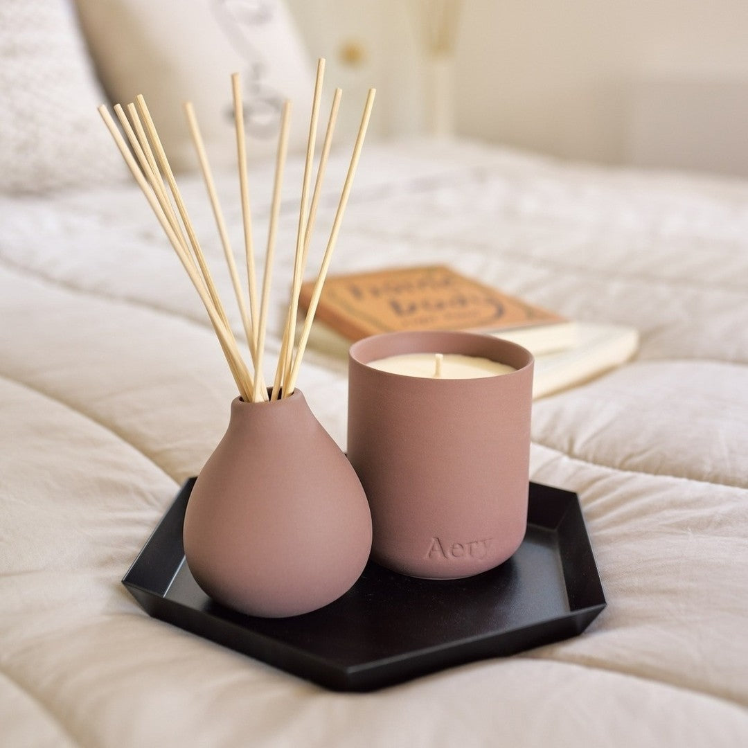 Earthy pink ceramic candle and diffuser on a black tray on a bed with light sheets